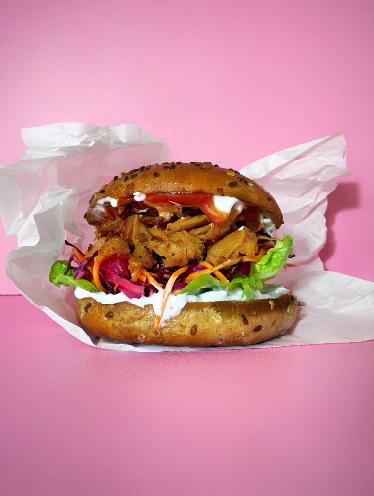 Vegan burger with chicken-style chunks and salad in a bap on white paper with a pick background